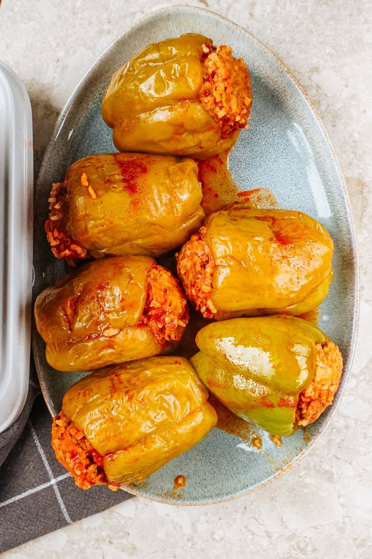 Six savory stuffed peppers, vibrantly green, cradle a flavorful red mixture. They're artfully arranged on a blue oval plate resting on a light marble surface.