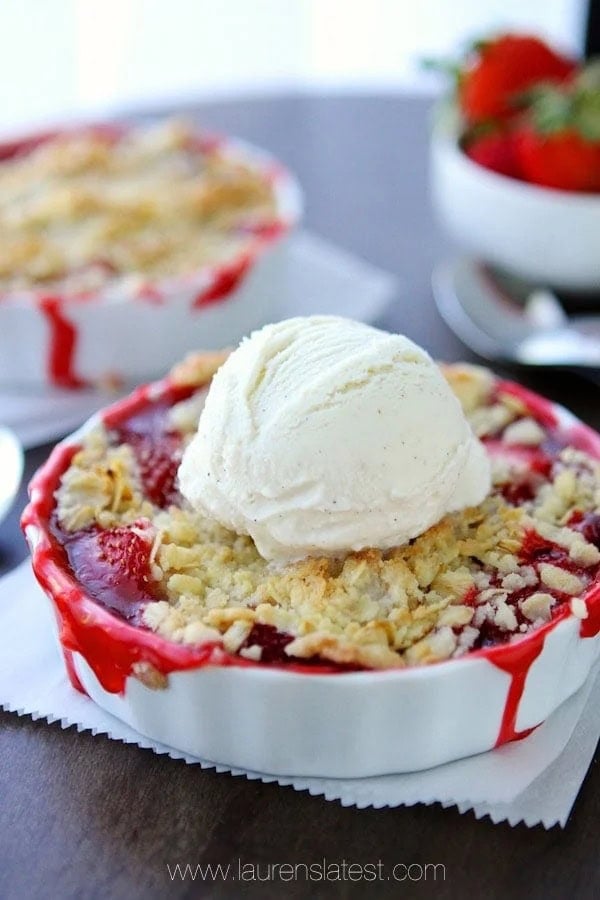 A strawberry crumble topped with a scoop of vanilla ice cream is in a white dish. A bowl of strawberries is blurred in the background.