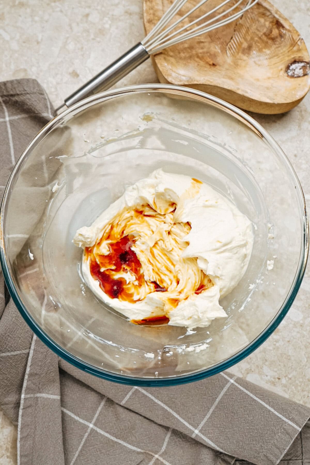 Glass bowl with cream cheese and hot sauce on a kitchen towel. Nearby are a whisk and a wooden bowl.