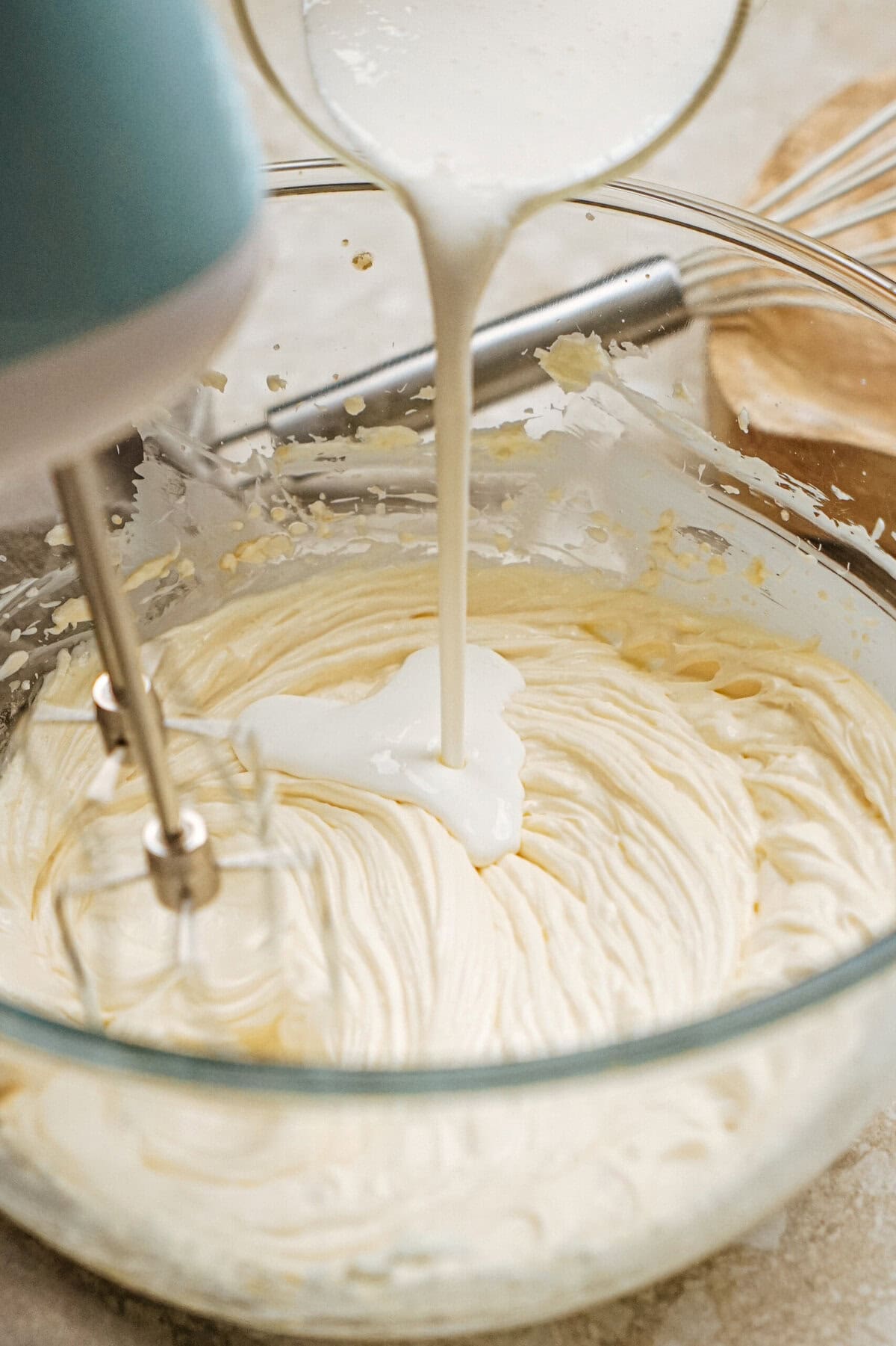 Electric mixer mixing cream being poured into thick batter in a glass bowl on a countertop.