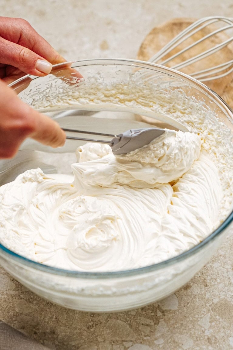 Person folding creamy white mixture in a glass bowl using a spatula, with a whisk and cutting board in the background.