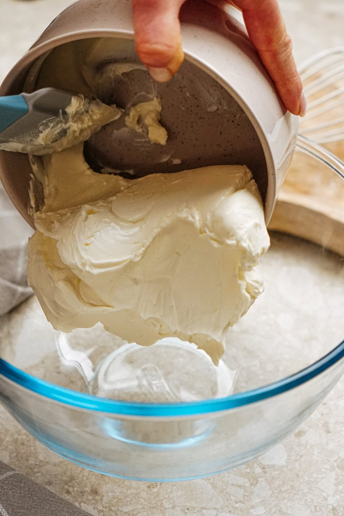 A person is scraping cream from a container into a glass bowl using a spatula.