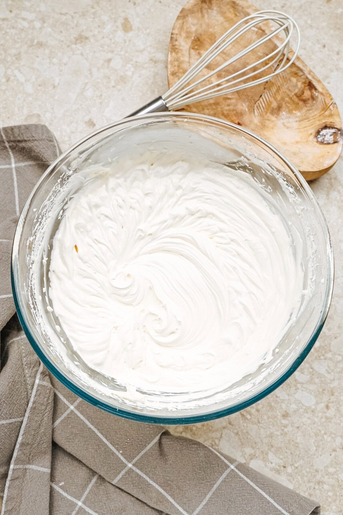 A glass bowl filled with whipped cream and a whisk on a wooden board, resting on a countertop next to a checkered cloth.