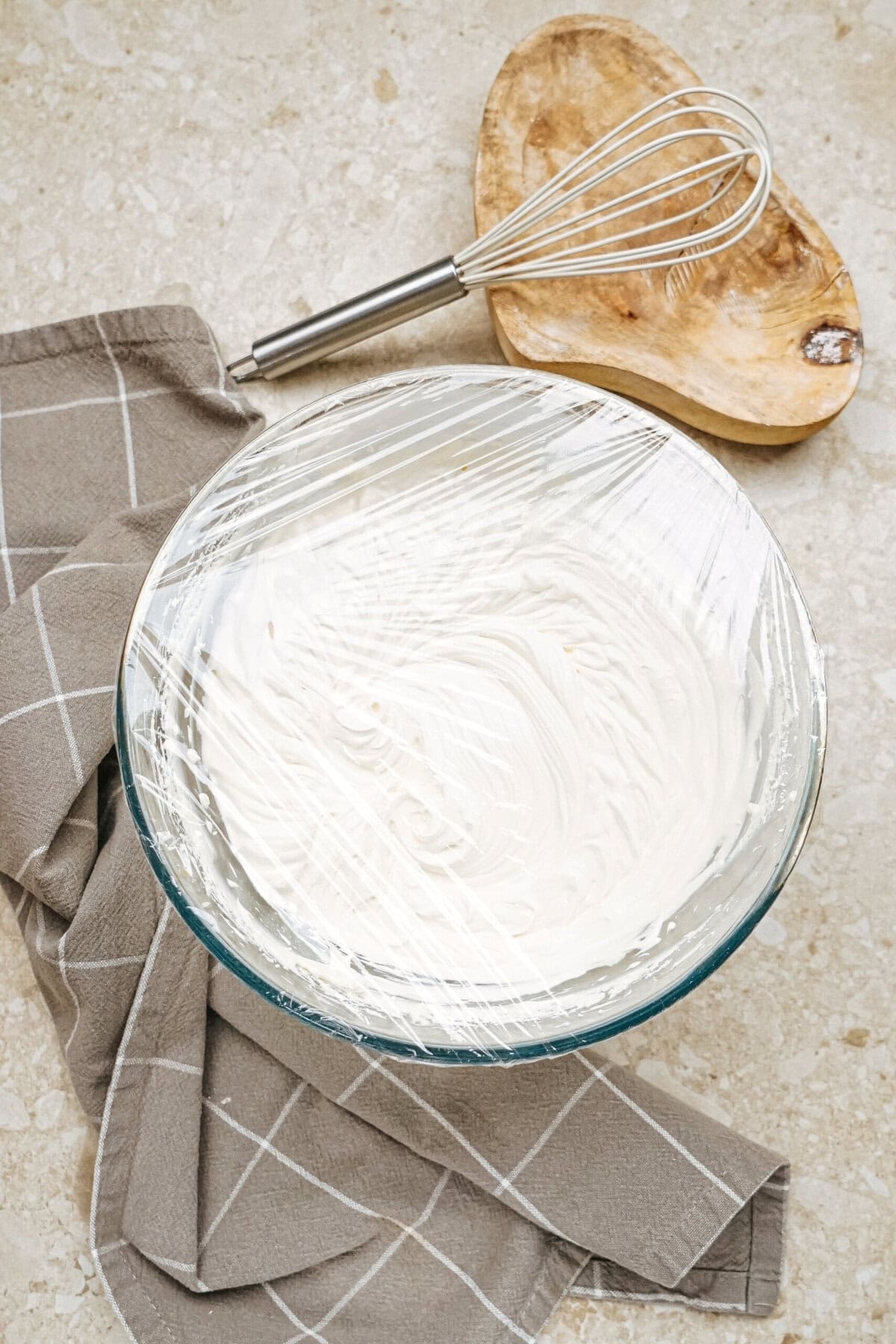 A glass bowl filled with cream, covered in plastic wrap, rests on a gray checkered towel. A wooden bowl and metal whisk are nearby on a light stone surface.