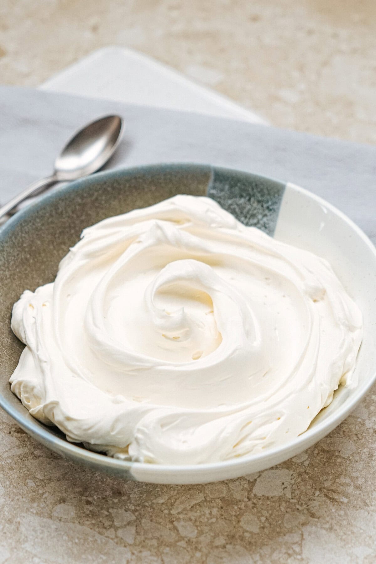 A bowl filled with smooth, swirled whipped cream on a beige surface, with a spoon placed beside it.