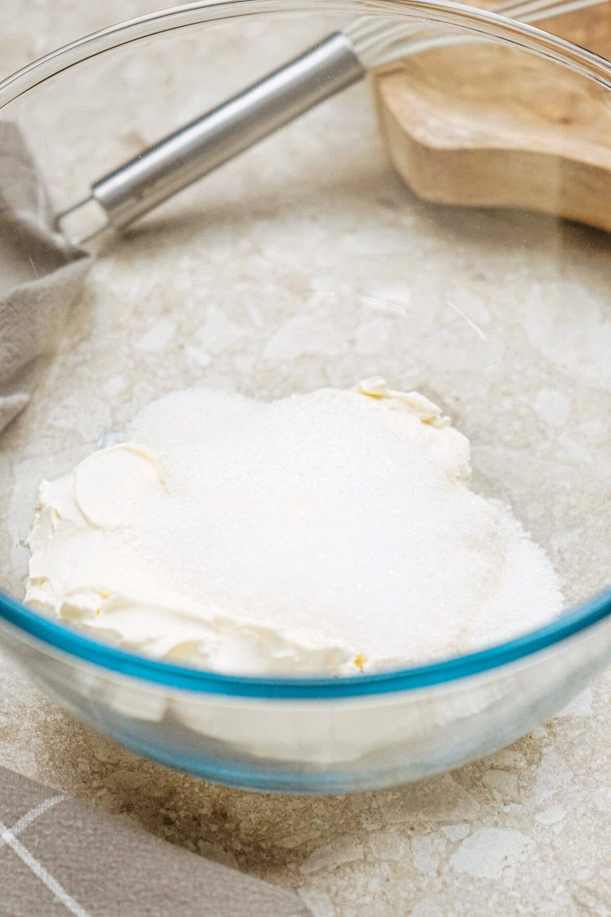 A glass bowl containing cream cheese and sugar on a countertop, with a whisk nearby.