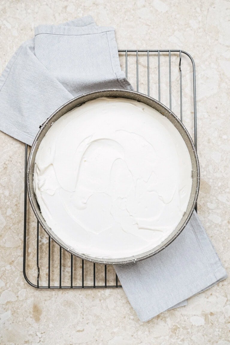 A round cake pan filled with smooth, white batter rests on a cooling rack, placed over a light gray cloth on a stone-textured surface.