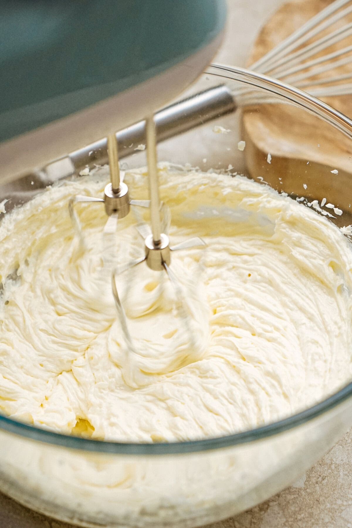 Electric mixer blending whipped cream in a glass bowl, with a whisk and wooden board in the background.