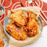 A plate of crispy fried chicken on parchment paper, with gold utensils and a red patterned napkin beside it on a marble surface.