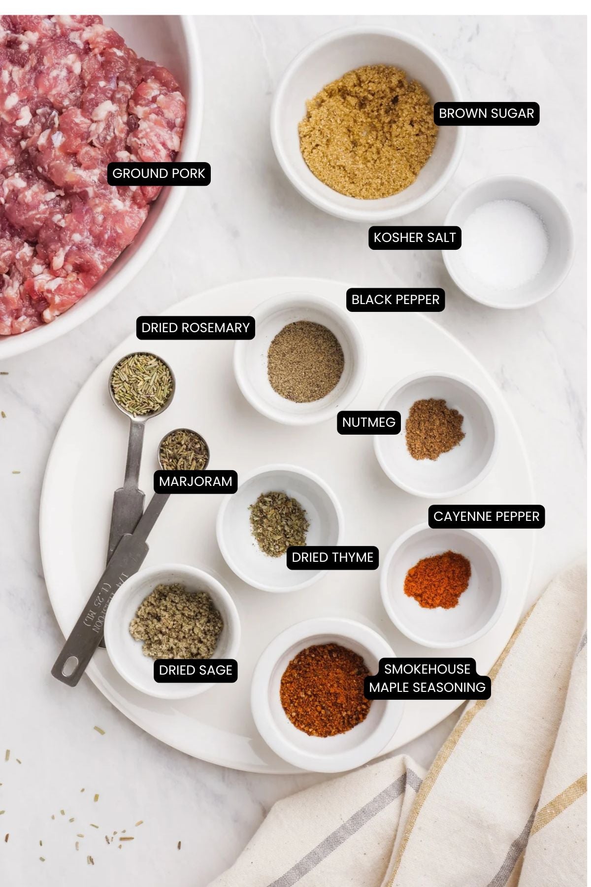 A plate holds nine small bowls and a spoon containing ground pork, brown sugar, kosher salt, black pepper, nutmeg, cayenne pepper, smokehouse maple seasoning, dried thyme, marjoram, rosemary, and sage for making breakfast sausage.