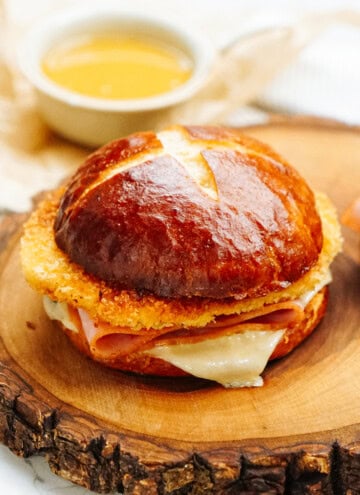 A sandwich with ham, melted cheese, and a crispy fried layer on a pretzel bun, served on a wooden board with a cup of dipping sauce in the background.