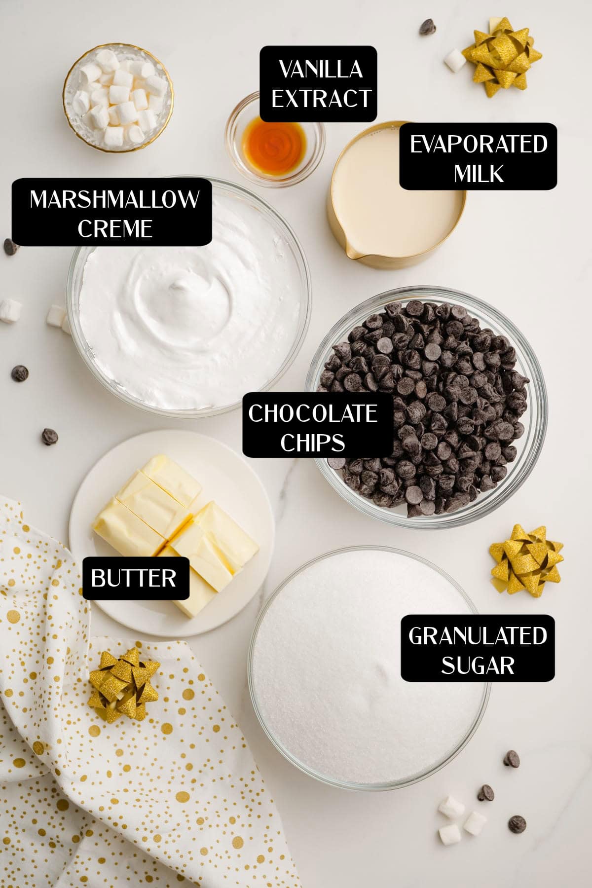Bowls and plates containing fudge ingredients like marshmallow creme, butter, granulated sugar, chocolate chips, evaporated milk, and vanilla extract are arranged on a white surface with decorative bows.