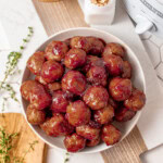 A bowl filled with glazed meatballs sits on a table near a crockpot, toothpicks, and sprigs of fresh thyme.