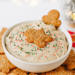 A bowl of creamy dip topped with festive sprinkles and gingerbread cookies, surrounded by assorted cookies on a platter.