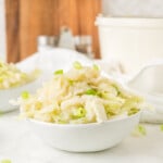 A white bowl filled with shredded cabbage salad garnished with chopped green onions, set on a white surface with kitchen items in the background.