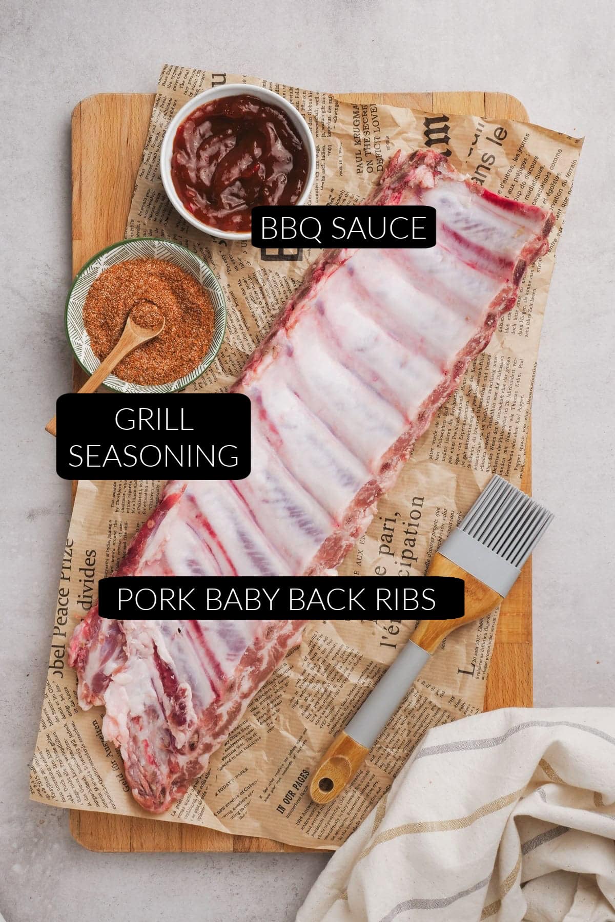A rack of raw pork baby back ribs in oven-ready form sits on parchment, with a bowl of grill seasoning, BBQ sauce, and a basting brush on a wooden board. Labels identify each item.