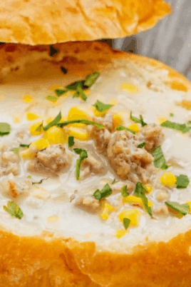 A bread bowl filled with creamy soup, garnished with chopped herbs and small pieces of meat, is placed on a grey plate.