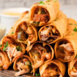 A stack of crispy rolled tacos filled with shredded chicken, diced vegetables, and melted cheese, garnished with chopped herbs on a wooden board.