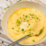 A bowl of mashed potatoes topped with chopped chives, black pepper, and a pat of butter, with a spoon in the bowl.
