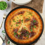 Golden baked pie topped with chopped parsley, surrounded by bowls of green peas and parsley, plates, and forks on a wooden board.