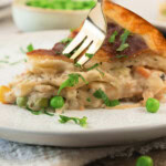 A fork is cutting into a slice of creamy turkey pot pie on a white plate, garnished with chopped parsley.