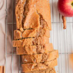 A loaf of apple bread, partially sliced, sits on a cooling rack with two whole apples, cinnamon sticks, and a small dish of sauce nearby.
