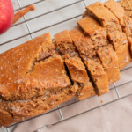 Sliced loaf of apple bread on a cooling rack with a red apple next to it, placed on a light-colored surface.