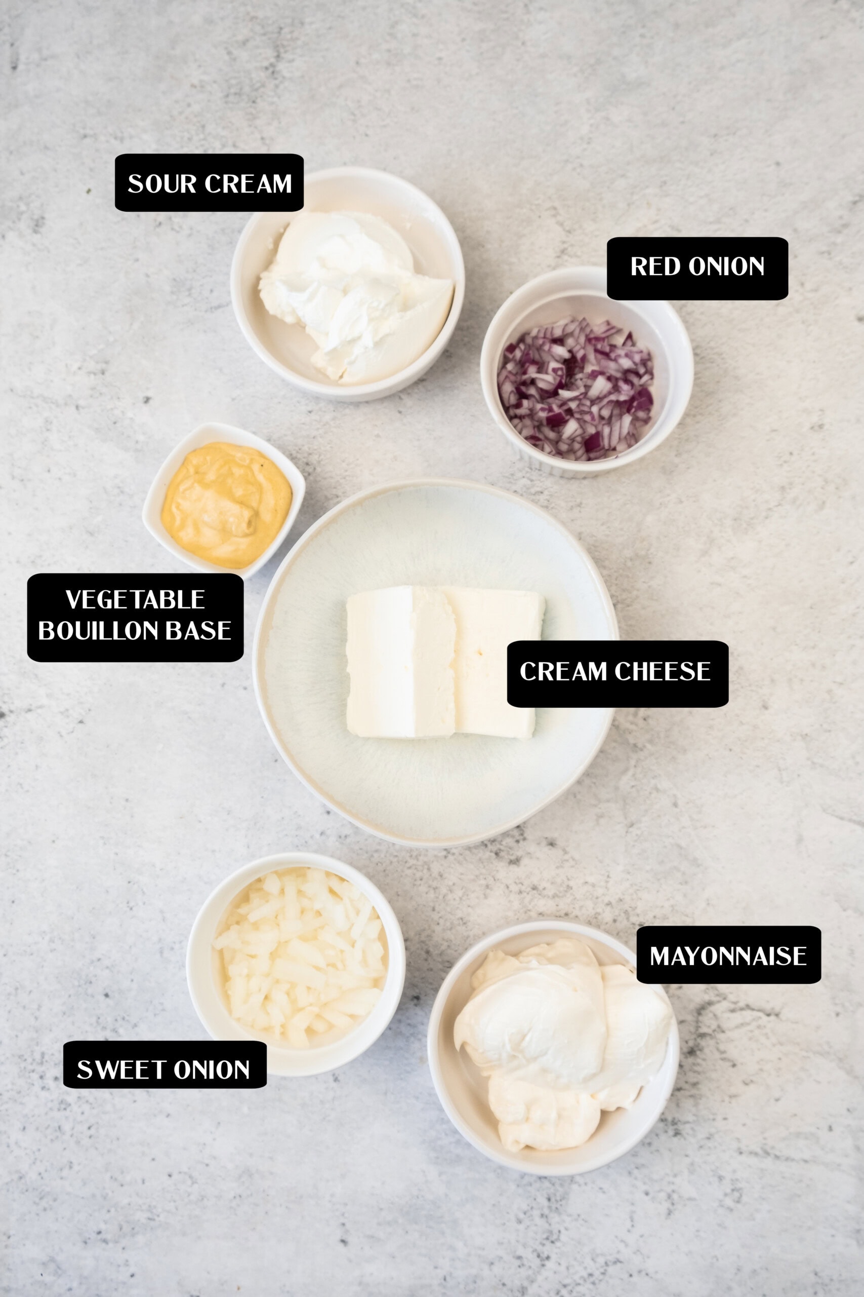 Six labeled bowls containing sour cream, red onion, vegetable bouillon base, cream cheese, sweet onion, and mayonnaise—perfect ingredients for making a classic onion dip—are arranged on a light surface.
