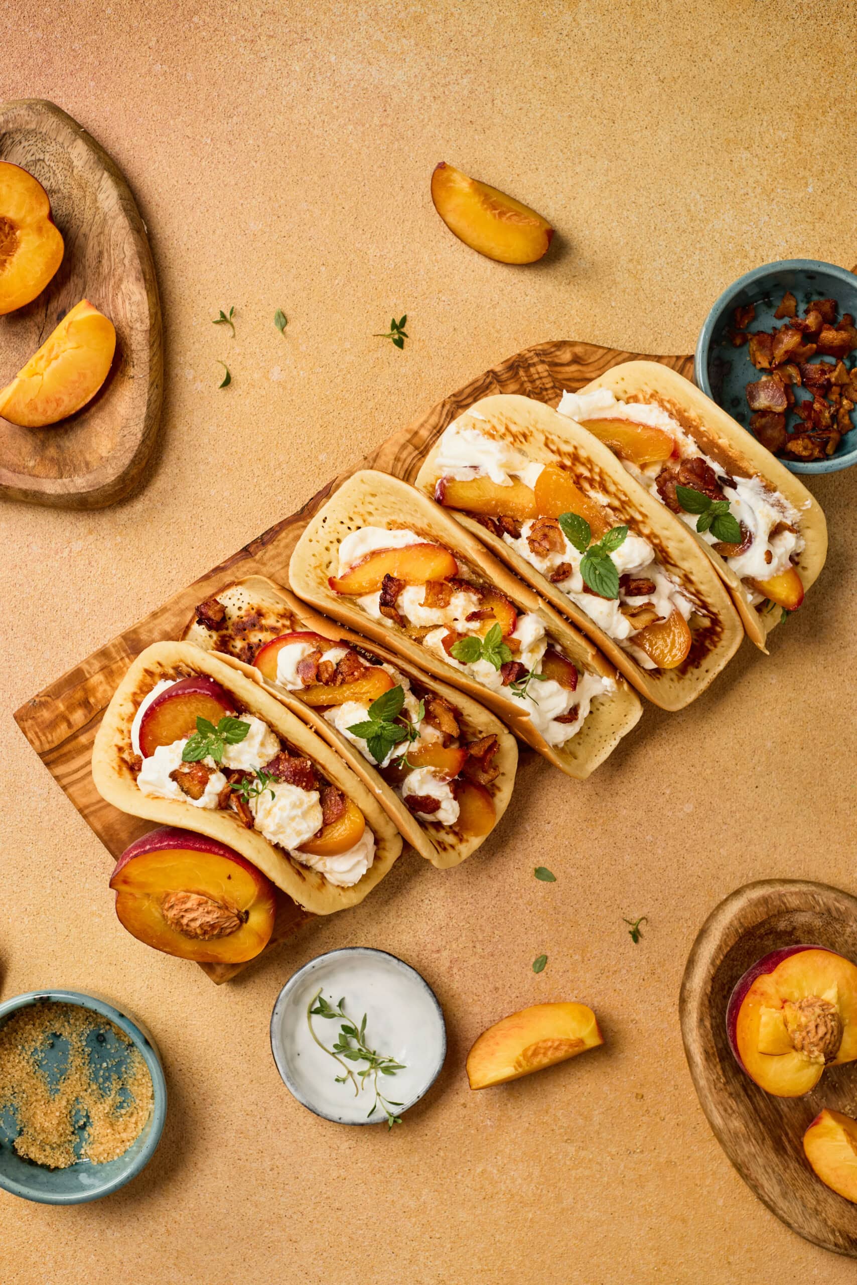 Four tacos filled with grilled chicken, peach slices, bacon, and a drizzle of sauce are arranged on a wooden board, surrounded by fresh peach wedges and small bowls of ingredients.