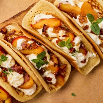 Three flatbread tacos filled with cream, grilled peaches, herbs, and bacon pieces, arranged on a wooden board with a bowl of white sauce and peach slices nearby.