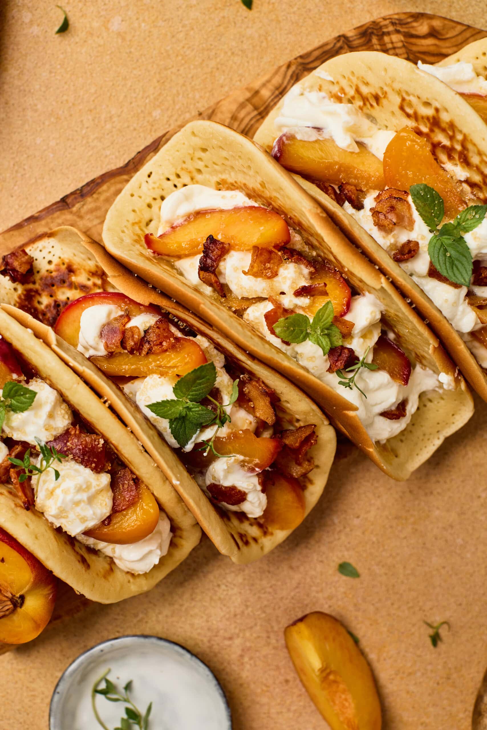 Three flatbread tacos filled with cream, grilled peaches, herbs, and bacon pieces, arranged on a wooden board with a bowl of white sauce and peach slices nearby.