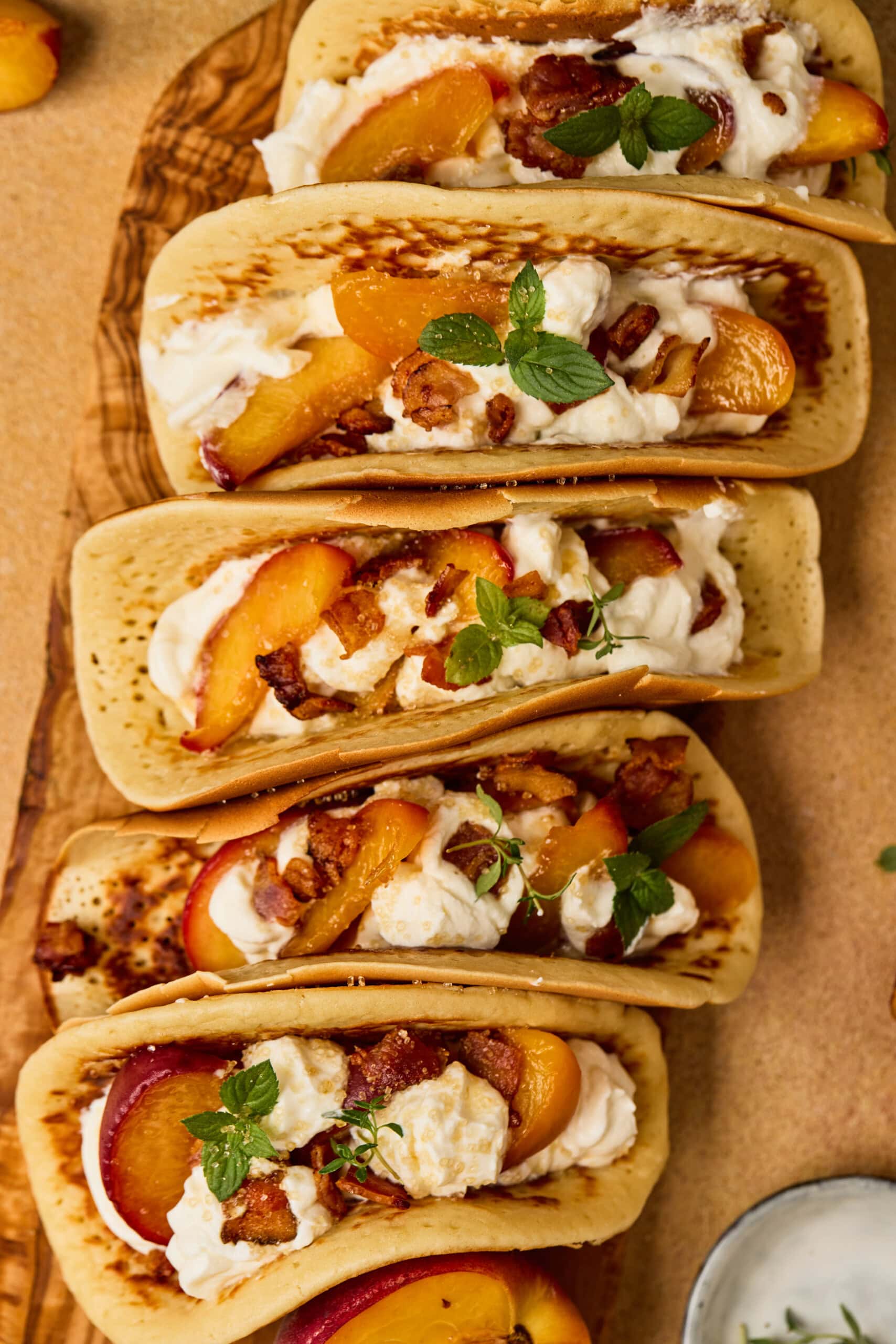 Five folded pancakes filled with whipped cream, peach slices, chopped pecans, and garnished with fresh mint leaves, arranged on a wooden board.
