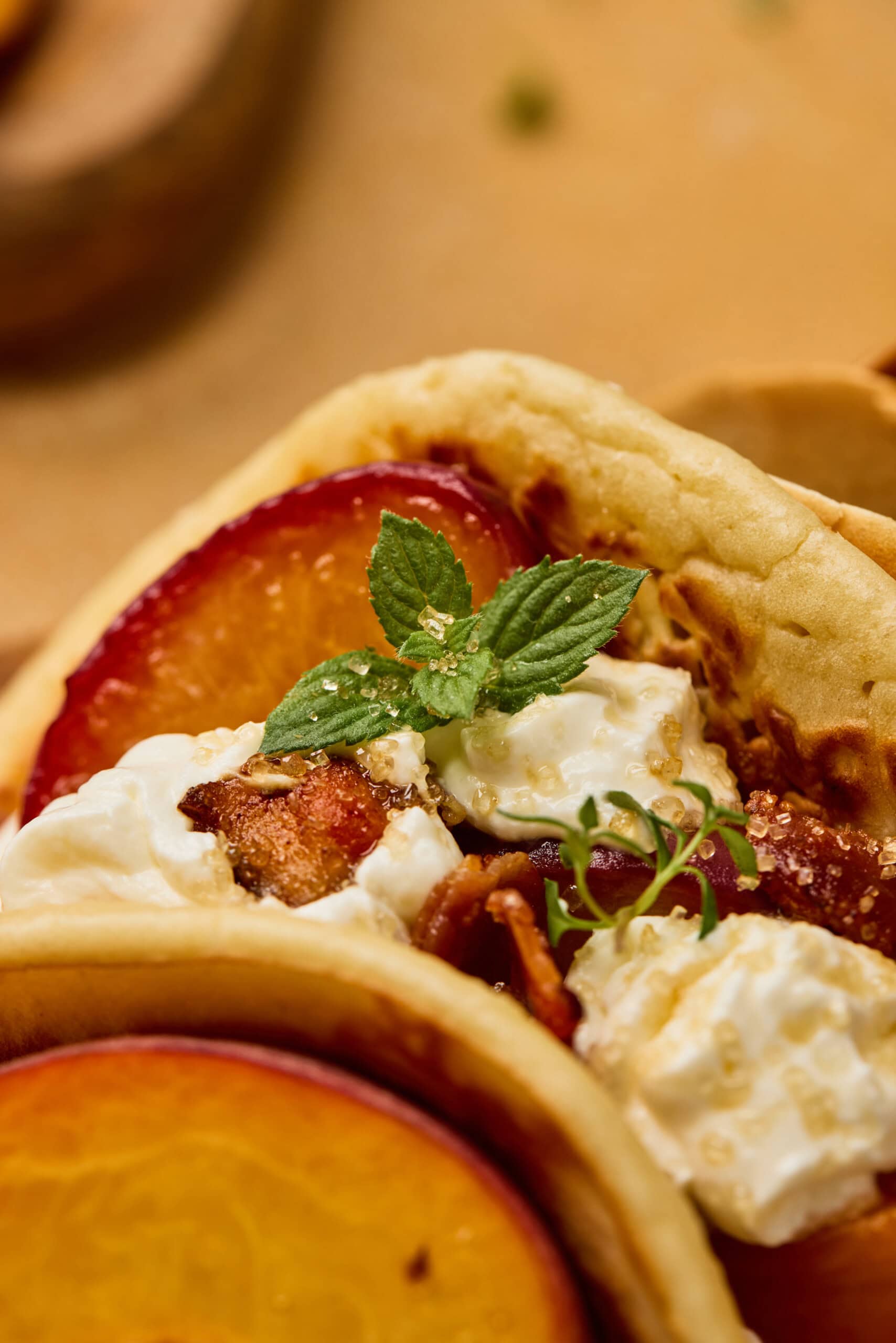 Close-up of a flatbread wrap filled with sliced peaches, bacon, whipped cheese, mint leaves, and a sprinkle of herbs.