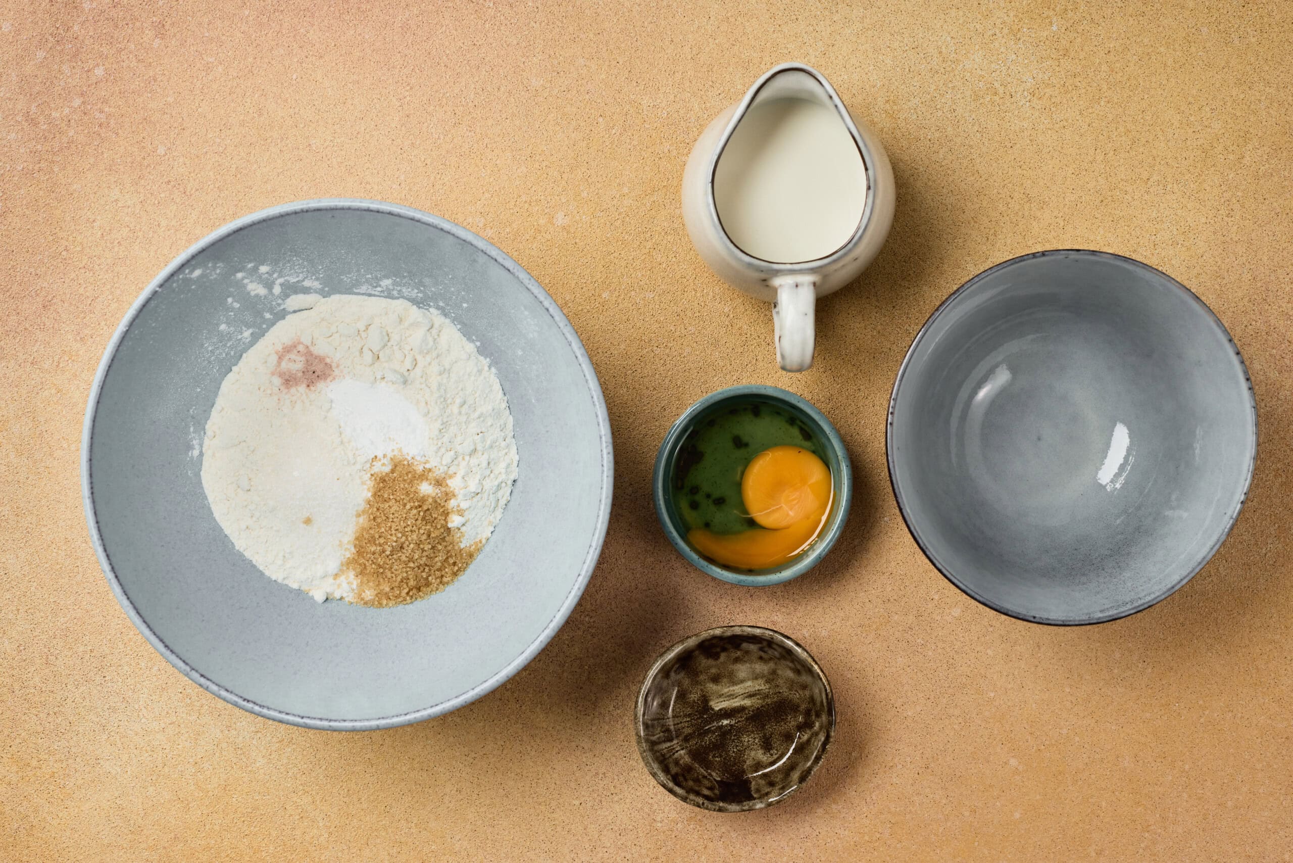 Five bowls on a beige surface containing flour and sugar, an egg, milk, an empty bowl, and a small bowl of another ingredient, arranged for baking preparation.