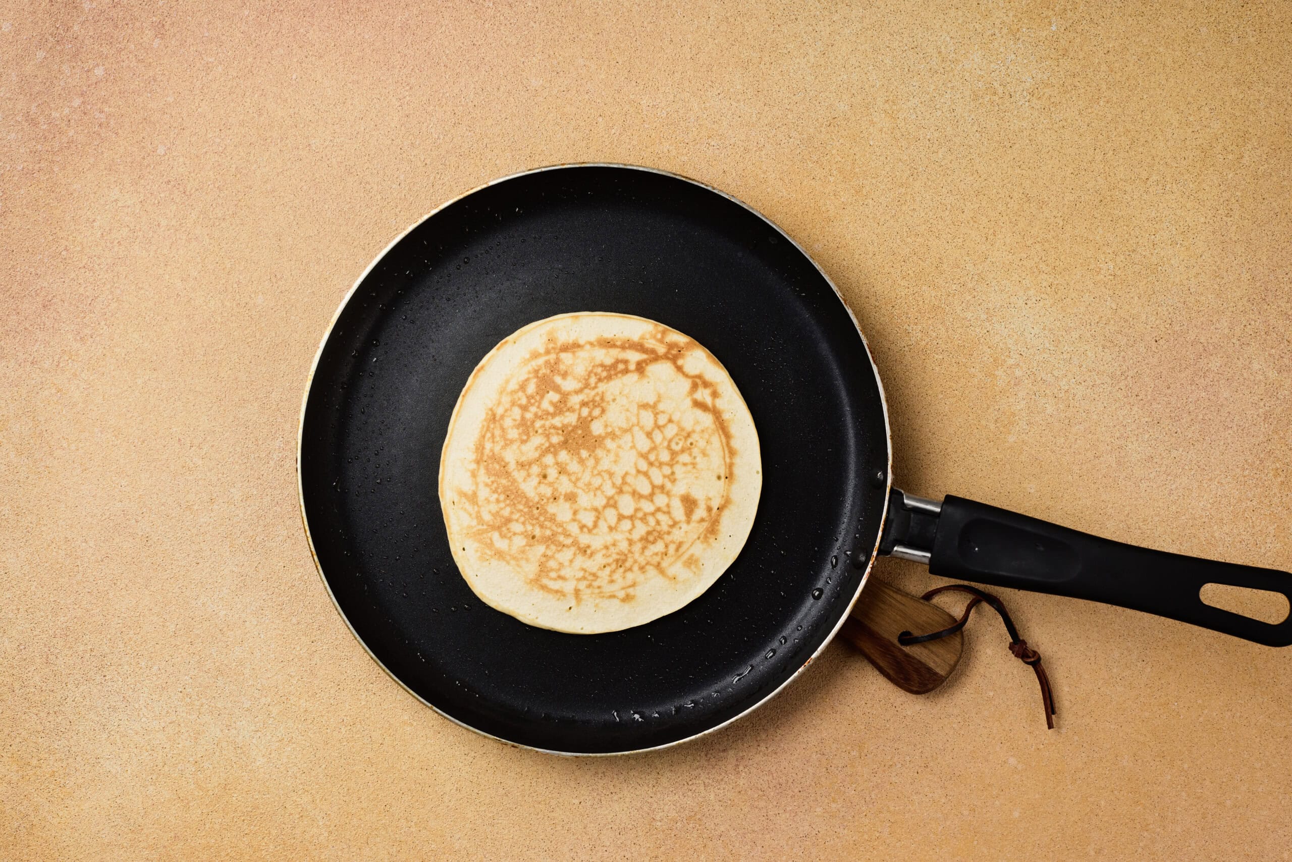 A single pancake cooking in a nonstick frying pan placed on a light brown countertop.