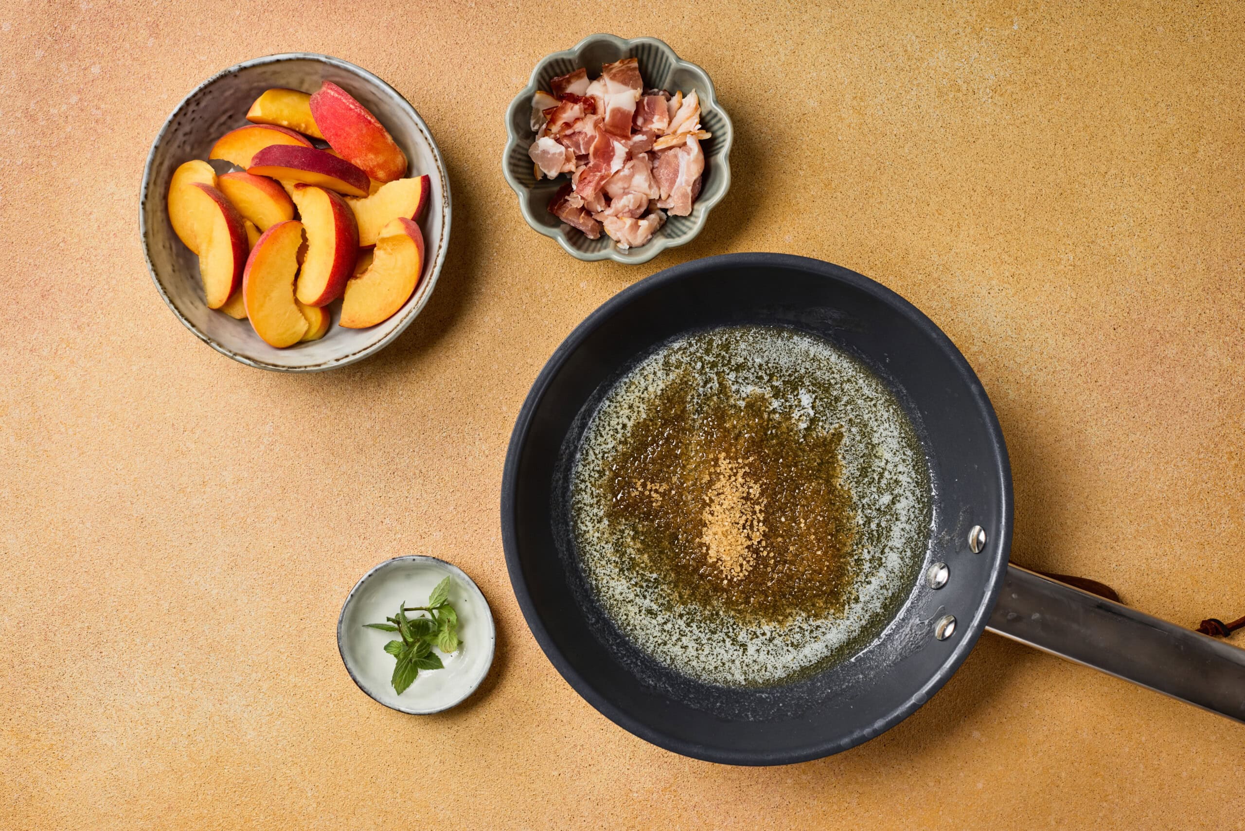 A frying pan with melted butter and brown sugar sits beside small bowls containing peach slices, chopped bacon, and fresh mint leaves on a beige surface.