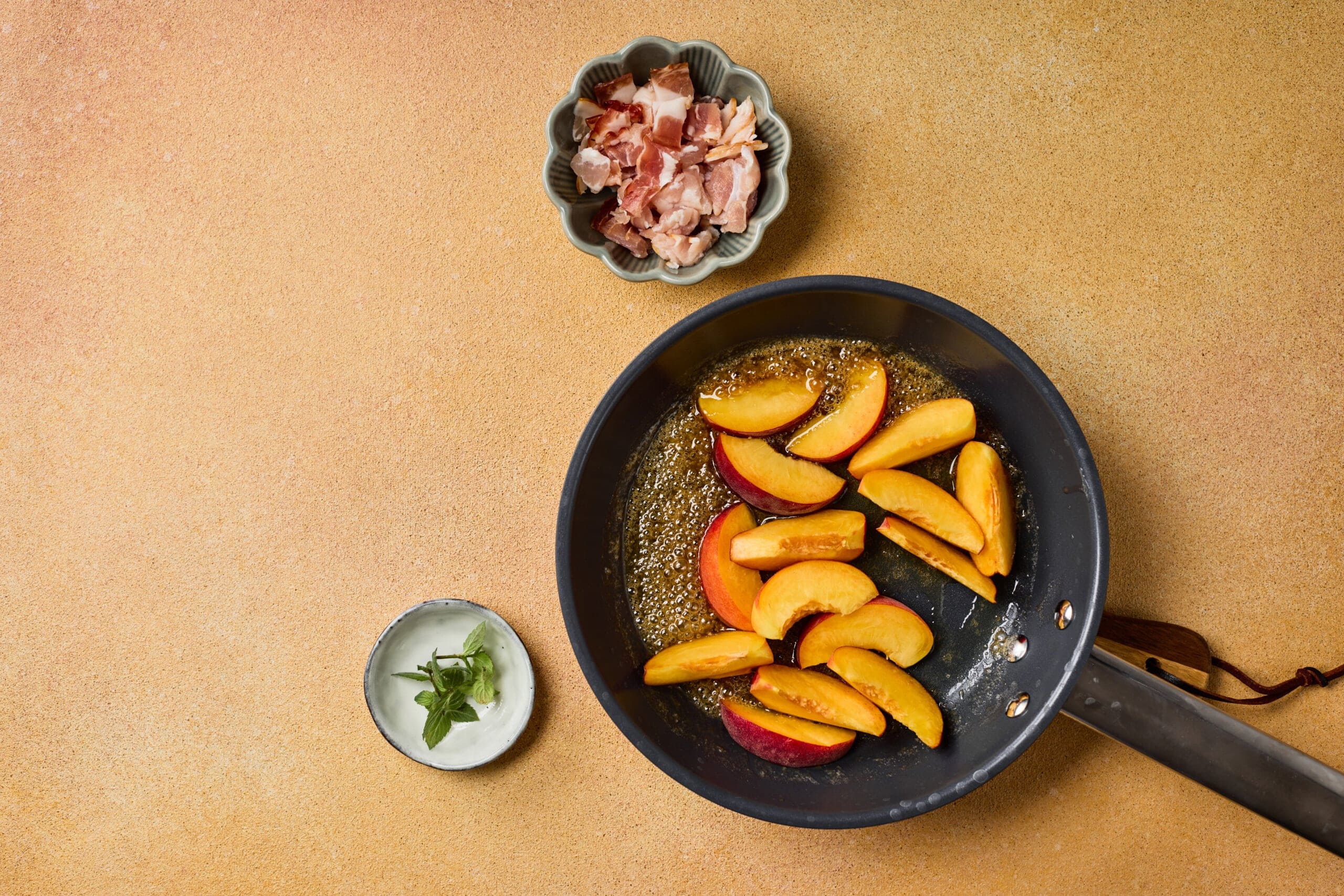 A frying pan with sliced peaches in syrup, a small bowl of chopped bacon, and a cup of mint leaves on a tan surface.