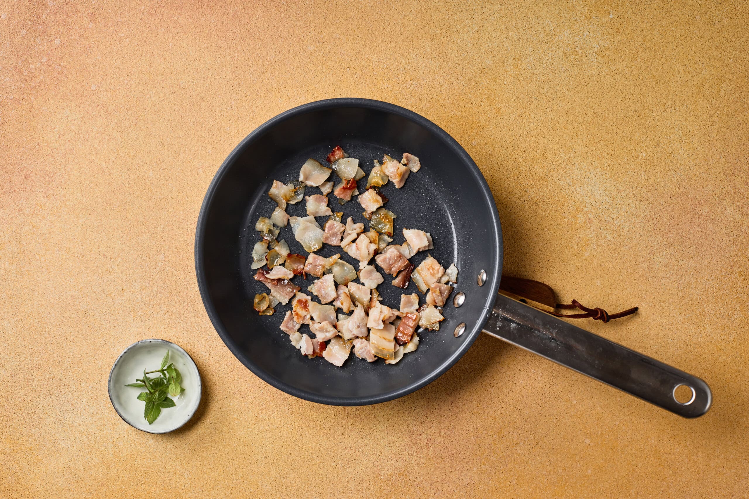 A frying pan with diced cooked meat and vegetables, placed on a tan surface next to a small bowl containing green herbs.