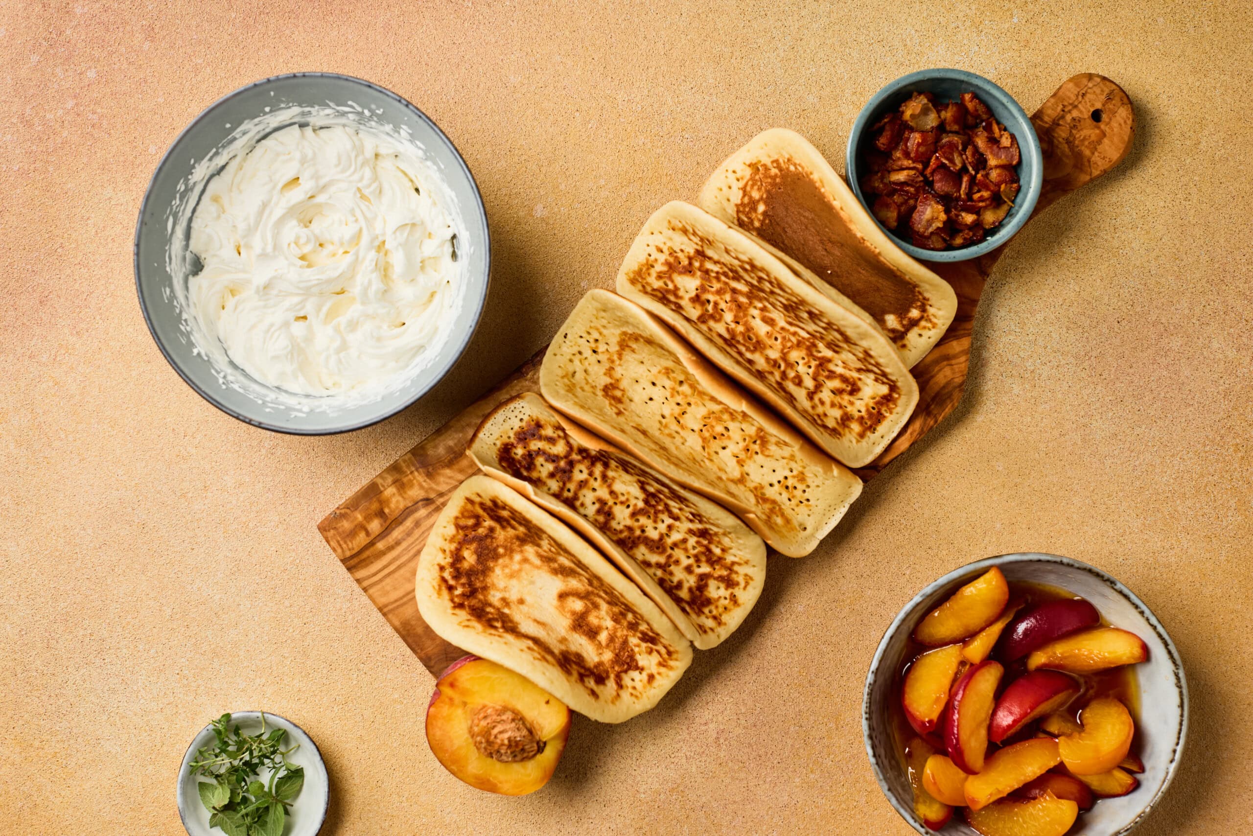 A wooden board with five folded pancakes, a bowl of whipped cream, a bowl of chopped nuts, a bowl of sliced peaches, a fresh peach, and a small bowl of herbs on a tan surface.