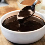 A spoon holds up thick chocolate sauce above a white bowl filled with chocolate sauce, with a bowl of cocoa powder in the background.