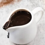A small white ceramic pitcher filled with thick chocolate sauce, with some sauce dripping down the spout, on a light marble surface.