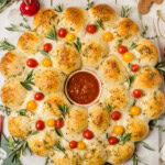 A round pull-apart bread wreath topped with cherry tomatoes and rosemary, arranged around a bowl of marinara sauce on a white wooden table with herbs and decorations.