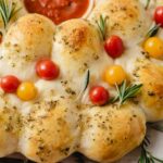 A wreath-shaped pull-apart bread topped with red and yellow cherry tomatoes, fresh rosemary, and herbs, with a bowl of marinara sauce in the center.