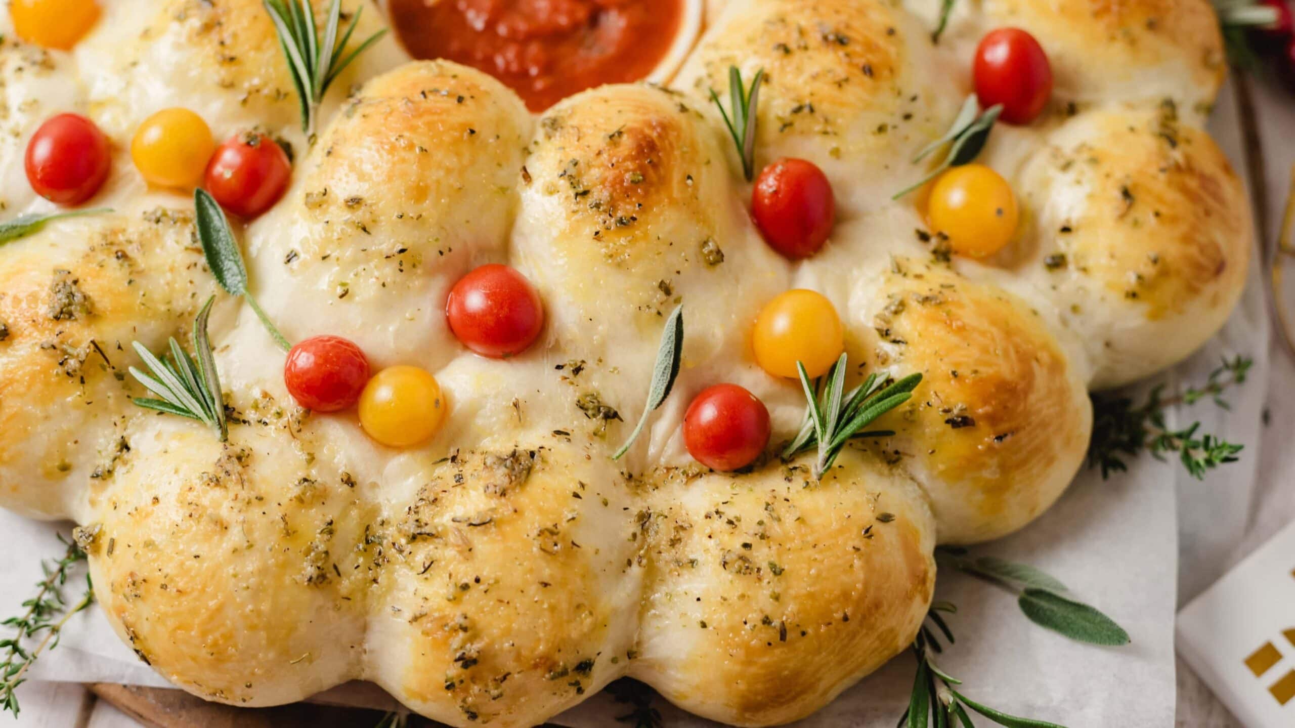 A wreath-shaped pull-apart bread topped with red and yellow cherry tomatoes, fresh rosemary, and herbs, with a bowl of marinara sauce in the center.