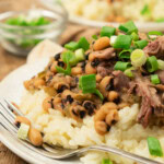 A plate of rice topped with black-eyed peas, shredded meat, and chopped green onions, with a fork on the side and a small bowl of green onions in the background.