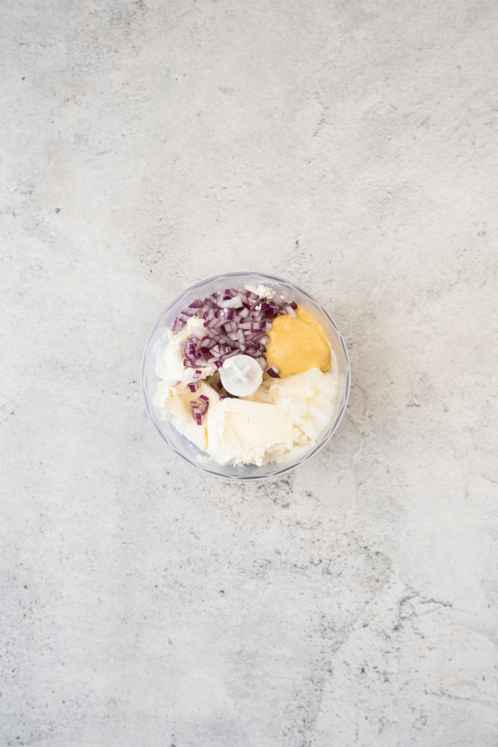 A food processor bowl containing chopped red onion, chopped white onion, cream cheese, and mustard on a light gray countertop.