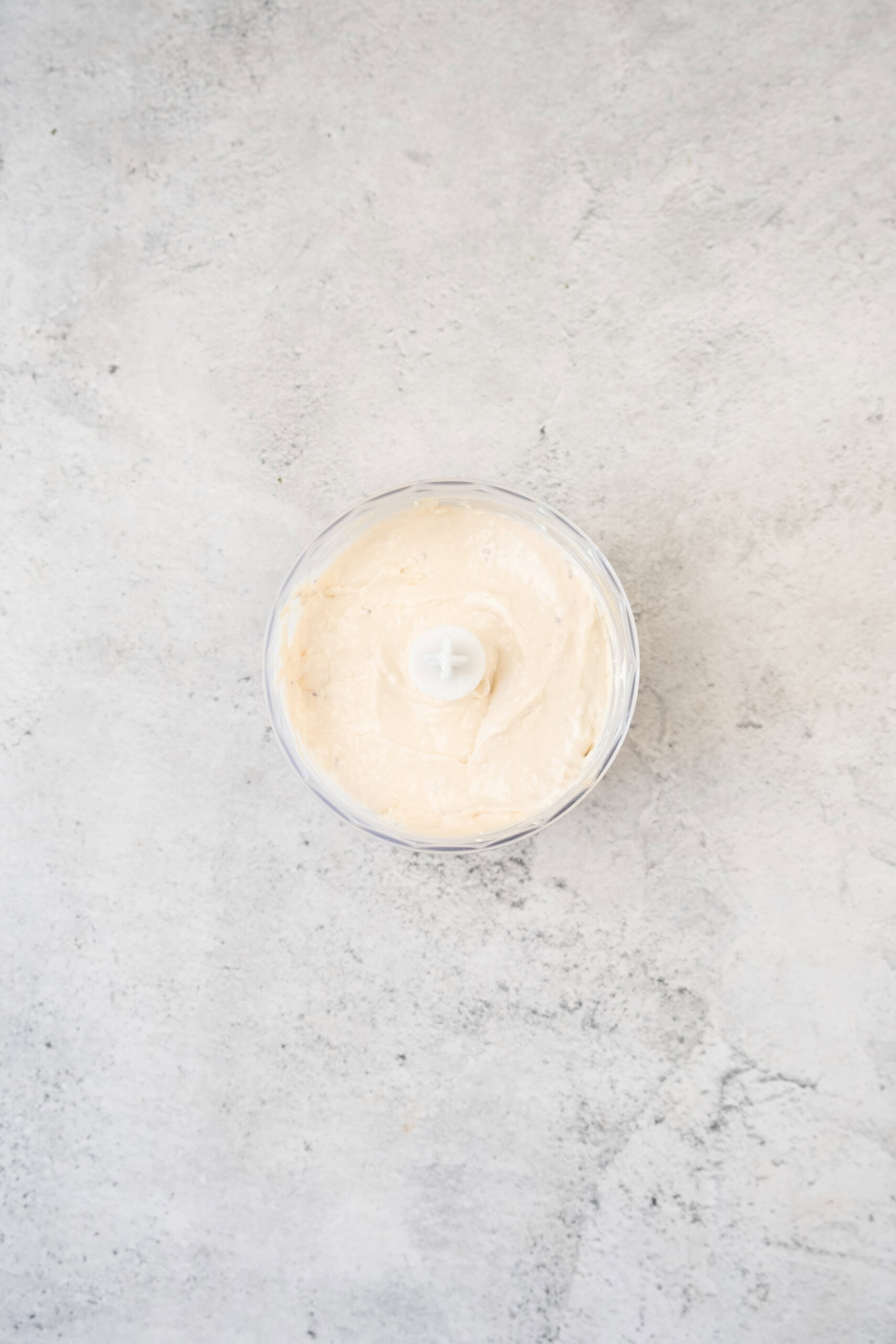 A small food processor bowl filled with smooth, creamy white spread sits on a light gray textured surface.