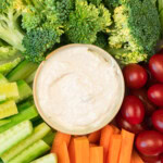 A bowl of white dip surrounded by fresh broccoli, cherry tomatoes, cucumber sticks, and carrot sticks.
