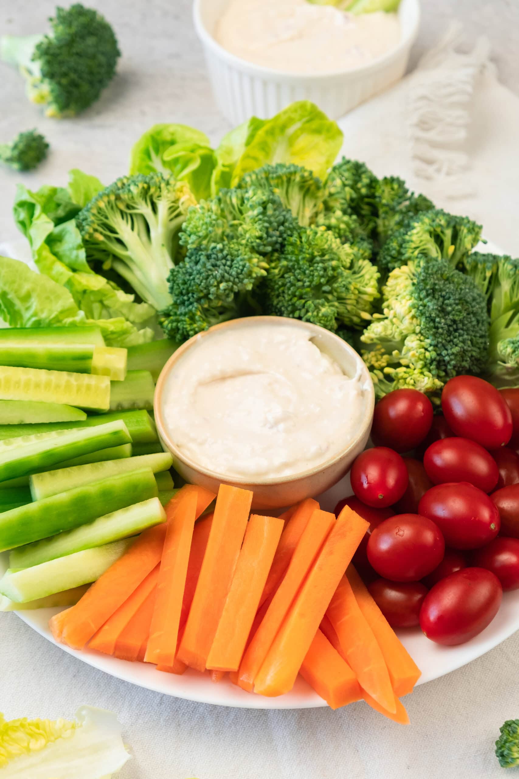 A white plate with broccoli florets, celery sticks, carrot sticks, grape tomatoes, and cucumber sticks arranged around a bowl of white dip.
