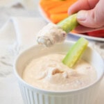 A hand dips a celery stick into a bowl of creamy dip, with a plate of assorted vegetables visible in the background.